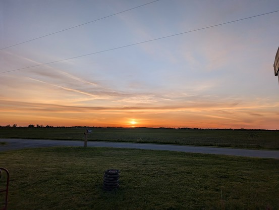 Sunrise over the corn field. Not today. The sun is just over the corn, and smeared into a multi part blob. The clouds around it are glowing bright orange, and helping the smeared effect by crossing parts of the sun at top and bottom. There's a line of gray and gold cloud that stretches almost all the way across the screen, and it's blue sky above that