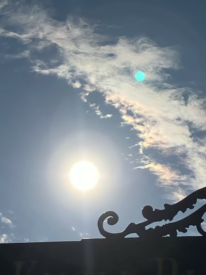 A bright sun, surrounded by wispy clouds against a blue background. The silhouette of an ornate iron street sign frames the bottom corner. 