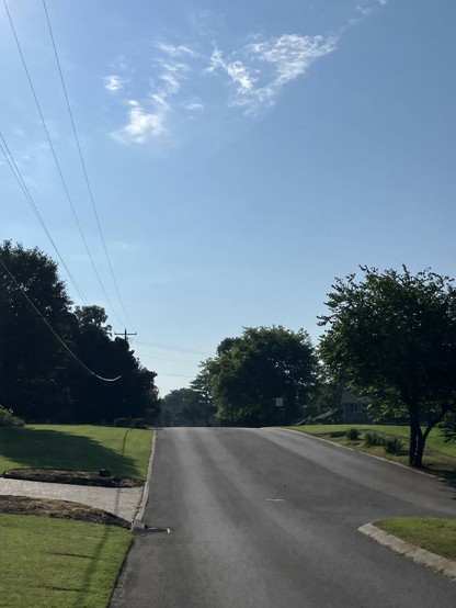 A view of a quiet residential street on a sunny day, featuring green grass and trees on either side of the road, which slopes gently upward. The sky is clear with a few clouds. Power lines are visible in the distance.