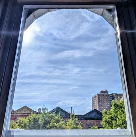 Looking through an arched window three hours after sunrise the light blue sky is full of amorphous windswept white clouds. The sun is just out of frame on the upper left, creating a subtle lens flare, a curved horizontal white line. Pointed roofs of Harlem brownstones with red brickwork are across the street, and a taller apartment building can be seen in the distance. The green tops of two trees are on the bottom and right.