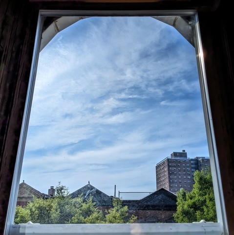 Looking through an arched window three hours after sunrise the blue sky is full of streaked white clouds. Pointed roofs of Harlem brownstones with red brickwork are across the street, and a taller apartment building can be seen in the distance. The green tops of two trees are on the bottom and right.