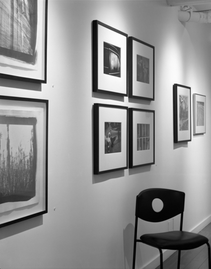 A black an white photo of photographs hanging in a gallery, close to us and partially out of frame are two pieces mounted above each other, followed by a group of four hung in a square, at the far end are a pair next to each other. A black chair is placed at an angle against the wall below the group of four. 