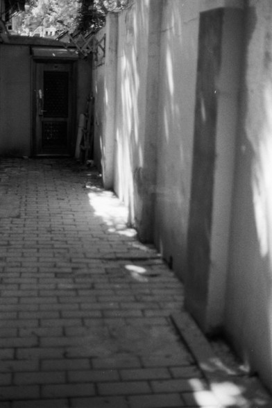 Fujifilm Neopan 100 SS (FF)

**English:**
A black and white photograph depicting a narrow alleyway with a brick-paved path. The alley is flanked by high walls on both sides, creating a sense of depth and leading the eye towards a door at the end. The shadows cast by the walls create a pattern of light and dark on the ground, adding texture to the scene. The door at the end of the alley is slightly ajar, with a ladder leaning against the wall nearby.

**Chinese:**
一张黑白照片，描绘了一条狭窄的小巷，铺着砖块的小路。小巷两侧是…