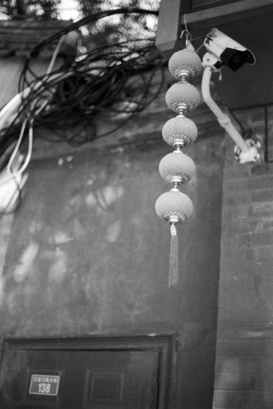 Fujifilm Neopan 100 SS (FF)

**English:**
A black and white photograph capturing a string of traditional Chinese lanterns hanging from a wire. The lanterns are intricately designed and appear to be made of a translucent material. They are positioned in front of a building with a numbered plaque, and a security camera is visible on the right side of the image. The lanterns add a cultural and festive element to the scene.

**Chinese:**
一张黑白照片，拍摄了一串悬挂在电线上的中国传统灯笼。灯笼设计精美，看起来是由半透明材料制成。它们位于一栋带有编号牌匾的建筑…