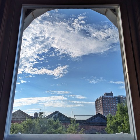 Looking through an arched window two hours after sunrise the bright blue sky is full of complex white clouds. Pointed roofs of Harlem brownstones are silhouetted across the street, and a taller apartment building can be seen in the distance. The green tops of two trees are on the bottom and right.