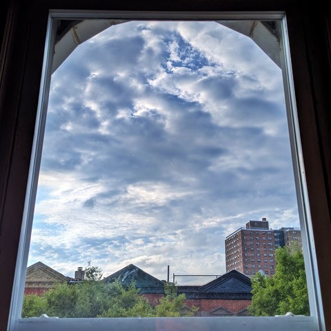 Looking through an arched window two hours after sunrise the sky is full of chunky white and blue clouds. Pointed roofs of Harlem brownstones with red brickwork are across the street, and a taller apartment building can be seen in the distance. The green tops of two trees are on the bottom and right.