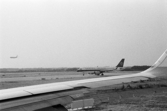 Foma FOMAPAN 400 Action (FF)

**English:**
This black and white photograph captures a view from an airplane window, showing the wing of the airplane extending into the frame. In the background, another airplane is visible on the runway, and a distant airplane is captured in mid-flight, adding a dynamic element to the scene. The landscape below appears to be an airport with grassy areas and some infrastructure. The image evokes a sense of motion and the vastness of air travel.

**Chinese:**
这张黑白…