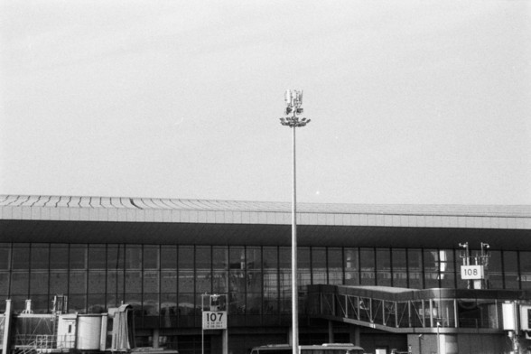 Foma FOMAPAN 400 Action (FF)

**English:**
This black and white image shows a section of an airport terminal building. The terminal features large windows and is marked with signs indicating gate numbers, such as "107" and "108." A tall light pole stands prominently in the foreground, adding to the composition of the photograph. The image conveys a sense of structure and organization typical of airport environments.

**Chinese:**
这张黑白照片展示了机场航站楼的一部分。航站楼带有大窗户，并标有登机口号码，如“107”和“108”。一根高高的灯杆矗立在前景，增添…