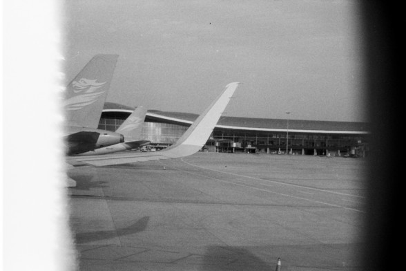 Foma FOMAPAN 400 Action (FF)

**English:**
This black and white photograph captures a view from an airplane window. The image prominently features the tail sections of two airplanes parked on the tarmac. The tails are angled towards each other, creating a sense of depth and perspective. In the background, an airport terminal building with large windows is visible, suggesting a busy airport environment. The overall scene evokes a sense of travel and aviation.

**Chinese:**
这张黑白照片是从飞机窗户拍摄的。图片中突出展…