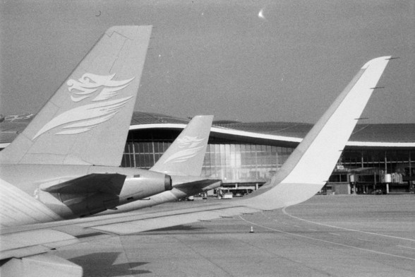 Foma FOMAPAN 400 Action (FF)

**English:**
This black and white image shows a closer view of the tails of three airplanes lined up on the tarmac. The tails are adorned with a distinctive logo, and the perspective emphasizes the alignment and symmetry of the aircraft. The airport terminal building is visible in the background, characterized by its large, reflective windows. The image conveys a sense of order and precision in the aviation industry.

**Chinese:**
这张黑白照片展示了跑道上排成一列的三架飞机的尾部特写。飞机尾部装饰着…