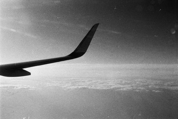 Foma FOMAPAN 400 Action (FF)

**English:**
A black and white photograph taken from an airplane window. The image captures a view of the airplane wing on the left side, extending outwards. Below the wing, there is a vast expanse of clouds with varying densities, creating a textured landscape. The horizon is faintly visible in the distance, separating the clouds from the sky, which appears smooth and layered.

**Chinese:**
一张从飞机窗户拍摄的黑白照片。照片中捕捉到飞机左侧机翼向外延伸的景象。机翼下方是广阔的云层，云层密度各异，形成了一种纹理丰富的景观。远处的地平线隐约…