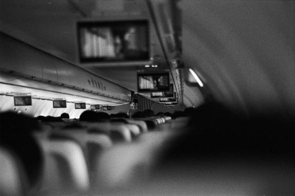 Foma FOMAPAN 400 Action (FF)

📝 English Alt Text
A black-and-white photo of an airplane cabin with passengers seated in rows. Overhead screens display in-flight entertainment, and a flight attendant stands in the aisle near the middle of the plane.

📝 中文替代文字
一张黑白色调的飞机客舱照片，乘客坐在整齐排列的座位上。天花板上挂着多个显示屏播放机上娱乐内容，一名空乘人员站在靠近客舱中部的过道中。