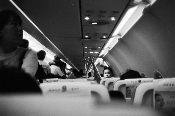 Foma FOMAPAN 400 Action (FF)

📝 English Alt Text
A black-and-white photo showing the interior of an airplane cabin, viewed from the rear. Passengers fill the seats and some are standing in the aisles, possibly preparing for takeoff or arrival. The scene captures a busy yet orderly atmosphere, with visible overhead compartments and soft cabin lighting.

📝 中文替代文字
一张从飞机尾部拍摄的黑白照片，展示了机舱内部。乘客坐满座位，有些人在过道上站立，似乎正在为起飞或到达做准备。画面呈现出繁忙但有序的氛围，行李舱和柔和的客舱灯光清晰可见。