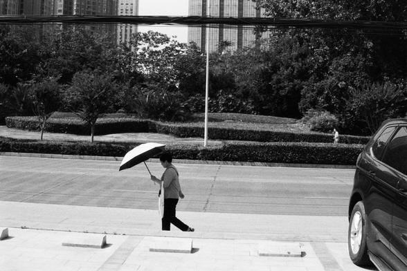 Foma FOMAPAN 400 Action (FF) ---

🔊 English (Screen Reader Friendly):
Monochrome photo of a person walking on a sidewalk while holding an umbrella. A car is parked on the right side of the road. In the background are neatly trimmed bushes, trees, and several tall buildings. The scene reflects a peaceful, rainy moment in a modern city environment.

🔊 中文（适合屏幕阅读器）：
黑白照片，一位行人打伞行走在人行道上。画面右侧是一辆停靠的汽车。背景是整齐的灌木、树木和高楼建筑。这幅图描绘了现代都市中雨天宁静的一刻。

---