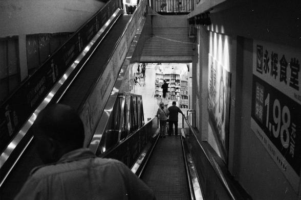 Foma FOMAPAN 400 Action (FF)

---
🔊 English (Screen Reader Friendly):
Black-and-white photo of an indoor area in China, featuring a set of escalators leading to a lower level. One person stands at the top, while two others descend together. Below is a store or supermarket with shelves stocked with products. Wall signs include large pricing in Chinese text—such as “1.98 yuan”—indicating promotional offers.

🔊 中文（适合屏幕阅读器）：
黑白照片，展示中国一处室内场景，中间是通往下层的自动扶梯。一人站在扶梯顶端，两人一起下行。下层是商店或超市，货架上摆满商品。墙上有中文标价，如“1.…