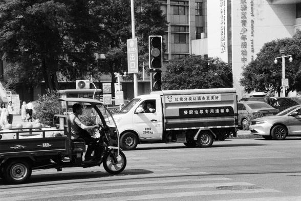 Foma FOMAPAN 400 Action (FF)

---

🔊 English (Screen Reader Friendly):
Black-and-white photo showing a busy urban street in China. A three-wheeled vehicle and a large kitchen waste transport truck are in the foreground. The truck bears Chinese text promoting trash sorting and urban cleanliness. Behind them, traffic lights, people, and tall buildings form a typical city backdrop.

🔊 中文（适合屏幕阅读器）：
一张黑白照片，展示中国城市街道。画面前景是一辆三轮车和一辆餐厨垃圾运输卡车。卡车上写有宣传垃圾分类和城市清洁的中文标语。背景中可见交通灯、行人以及高楼林立的城市景观。