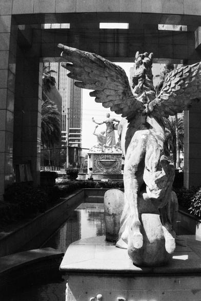 Foma FOMAPAN 400 Action (FF)

🇬🇧 English Alt Text:
A black and white photograph featuring a majestic winged horse statue, likely representing Pegasus, standing atop a fountain. Behind it, a separate statue of a human figure raises a torch or staff high into the air. The setting blends classical architecture—arched structures and columns—with towering buildings and palm trees in the distance, creating an intriguing contrast between mythological symbolism and modern urban surroundings.

🇨🇳 中文替代文本…