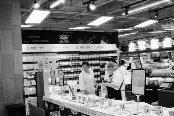 Foma FOMAPAN 400 Action (FF)

📝 English Alt Text

A black and white photo of a supermarket. At the food counter, a store employee wearing a hairnet and apron interacts with a customer. The counter displays various food items, and behind them, shelves filled with products and price signs are visible.

📝 中文替代文字（Alt Text）

一张黑白照片，场景为超市。在食品柜台前，一位戴着发网和围裙的店员正在与顾客互动。柜台上展示着各种食品，背景中的货架上摆满了商品，并有显示价格的标志。