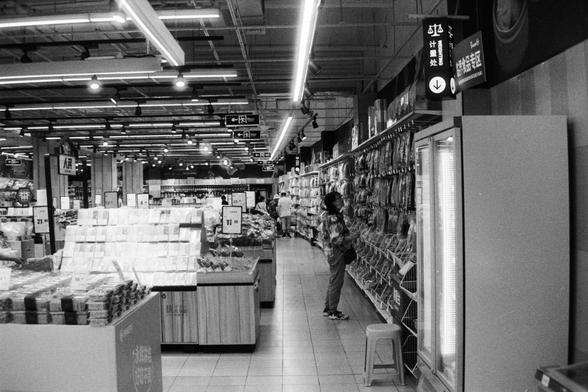 Foma FOMAPAN 400 Action (FF)

📝 English Alt Text

A black and white photo of a grocery store interior. A customer stands in an aisle filled with shelves stocked with packaged goods. Stacks of products with price tags are displayed in the middle of the store. Overhead, lights illuminate the space, and large hanging signs in Chinese indicate different sections. The overall atmosphere evokes a classic or documentary-style feel.

📝 中文替代文字（Alt Text）

一张黑白照片，显示一家杂货店的内部。顾客站在货架之间，货架上摆满了各类包装商品。商店中央有产品堆叠…