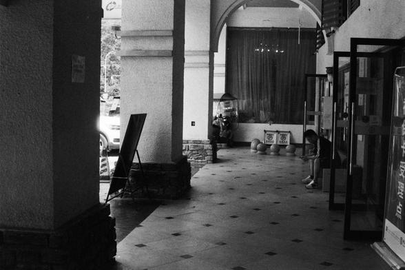 Foma FOMAPAN 400 Action (FF)

📝 English Alt Text

A black and white photo of a corridor with tiled diamond-pattern flooring. On the right, a person is seated on a bench near a glass door. Several decorative spherical objects are placed along the floor. The corridor features tall columns and arches, with a large curtained window and a display case at the far end. A car is partially visible outside. A standing signboard is placed near the entrance, contributing to the architectural elegance of th…