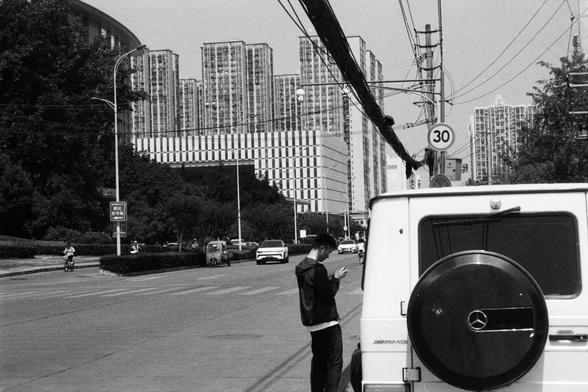Foma FOMAPAN 400 Action (FF)

📝 English alt text:
A black and white photo of an urban street scene featuring tall apartment buildings. A person stands beside a white Mercedes-Benz G-Class SUV, looking down at their phone. The road includes bicycles and cars, and overhead, power lines stretch above a speed limit sign reading 30 km/h.

📝 中文替代文本:
一张黑白城市街景照片，画面中有高层公寓楼。一人站在白色奔驰G级SUV旁边，低头看手机。道路上有自行车和汽车，上方是电线和限速30公里/小时的交通标志。