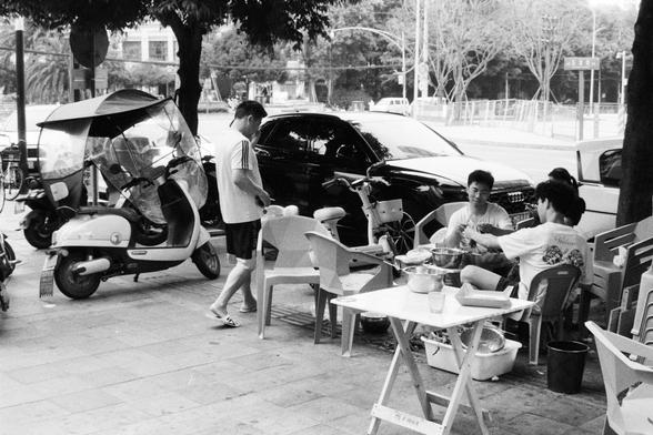 Foma FOMAPAN 400 Action (FF)

📝 English alt text:
A casual outdoor gathering scene featuring three people seated around a small table with bowls, containers, and miscellaneous items on it. A fourth person stands nearby, engaging with the group. To the left, a scooter covered with a tarp and a black car are parked along the street. The setting includes plastic chairs, surrounding trees, and distant buildings, suggesting a laid-back street-side atmosphere.

📝 中文替代文本：
一个休闲的户外场景，三人围坐在小桌旁，桌上放有碗、容器及其…