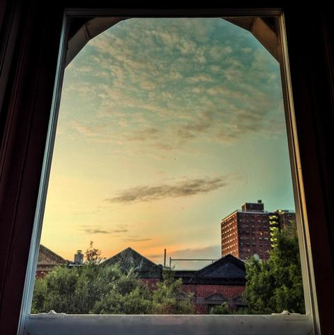 Looking through an arched window twelve minutes before sunrise the sky is cyan with a smear of orange above the horizon. The upper sky has flocked white clouds, and a long gray cloud is below. Pointed roofs of Harlem brownstones with red brickwork are across the street, and a taller apartment building can be seen in the distance. The green tops of two trees are on the bottom and right.