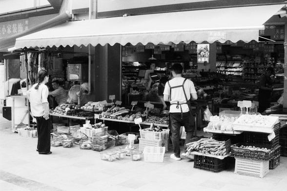 Foma FOMAPAN 400 Action (FF)

English Alt Text:
A black and white photo showing a busy market stall with baskets and crates full of fresh vegetables like carrots and cucumbers. Two people are seen—one standing and one working behind the stall—surrounded by produce both inside and outside the store.

中文替代文字（Chinese Alt Text）：
一张黑白照片，描绘一个繁忙的市场摊位，篮子和箱子里装满了胡萝卜、黄瓜等新鲜蔬菜。画面中有两个人，一个站着，一个在摊位后方工作，摊位周围和店内都堆满了农产品。
