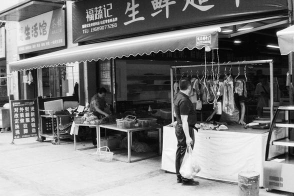 Foma FOMAPAN 400 Action (FF)

English Alt Text:
A street market scene featuring a meat shop with whole cuts of meat hanging on display. A man stands in front of the shop holding a grocery bag, observing the selection. Behind him, a woman arranges fresh produce at a table. The storefront displays Chinese signage with contact information and promotional text.

中文替代文字（Chinese Alt Text）：
街头市场场景，画面中心是一家肉铺，挂着整块肉类供顾客挑选。一名男子站在店前，手提购物袋，注视着展示的肉品。他身后是一位在桌旁整理蔬菜的女子。店面张贴有中文招牌和电话号码，以及宣传标语。