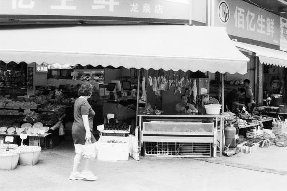 Foma FOMAPAN 400 Action (FF)

English Alt Text:
A bustling outdoor market scene with a variety of produce displayed in baskets and crates. Fresh fruits, vegetables, and meats hang from stalls, while a shopper walks by carrying a plastic bag. Several people are visible in the background engaging with the vendors. Above the stalls, a large awning features Chinese characters, indicating the name of the shop and contact details.

中文替代文字（Chinese Alt Text）：
一幅热闹的露天市场画面，篮子和箱子里摆满了各式水果、蔬菜和肉类。摊位上还挂着肉品，一位…