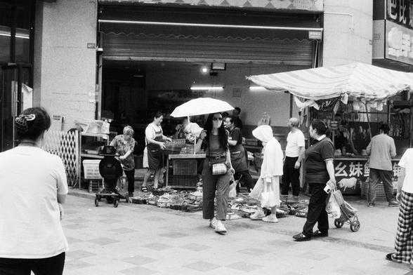 Foma FOMAPAN 400 Action (FF)

English Alt Text:
A busy street market scene captured. Several people are walking among stalls displaying various goods, with some merchandise placed directly on the ground or on tables. One person holds an umbrella while shopping. Others are seen standing or browsing items under coverings. The overall vibe is bustling yet cozy, as locals engage in market activity despite the weather.

中文替代文字（Chinese Alt Text）：
一张街头市场照片，多个摊位展示各种商品，有些物品摆放在地面或桌子上。一位顾客撑伞行走，其他人则在遮棚下站立或…