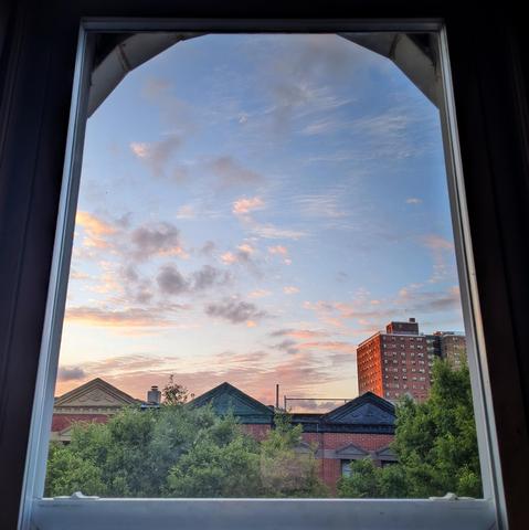 Looking through an arched window seven minutes after sunrise the sky fades from white at the horizon to baby blue up above, with scattered gray and pink clouds. Pointed roofs of Harlem brownstones with red brickwork are across the street, and a taller apartment building can be seen in the distance. The green tops of two trees are on the bottom and right. 