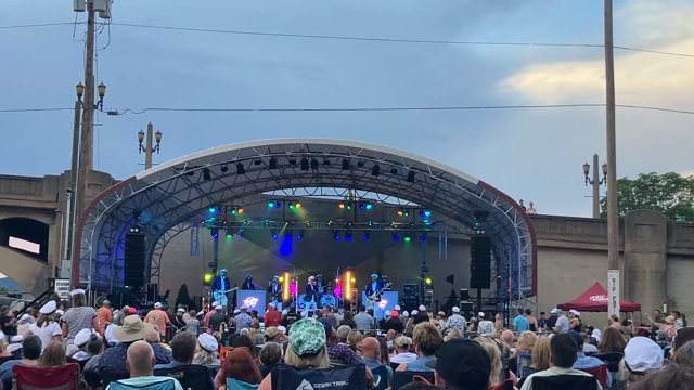 An outdoor concert pavilion. On stage, musicians wearing white sailor uniforms are playing Robbie Dupree’s “Steal Away”
