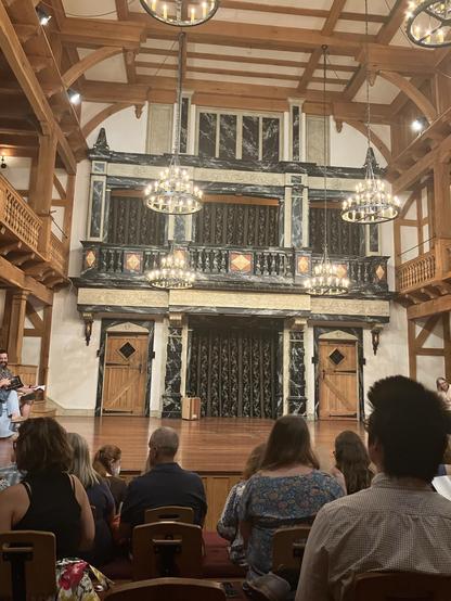 An audience is seated in a theater with a wooden interior. The stage features ornate decorations and large chandeliers. There are two wooden doors with diamond-shaped windows and a red and black patterned curtain in the background.
