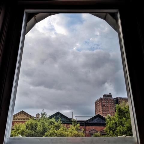 Looking through an arched window forty minutes after sunrise small patches of blue sky are visible behind thick gray and white clouds. Pointed roofs of Harlem brownstones with red brickwork are across the street, and a taller apartment building can be seen in the distance. The green tops of two trees are on the bottom and right.