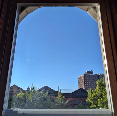 Looking through an arched window ninety minutes after sunrise the sky is bright blue and cloudless. Pointed roofs of Harlem brownstones are silhouetted across the street, and a taller apartment building can be seen in the distance. The green tops of two trees are on the bottom and right. 