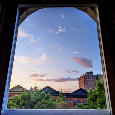 Looking through an arched window seven minutes after sunrise the sky fades from white at the horizon to deep blue up above, with scattered gray and white clouds. Pointed roofs of Harlem brownstones with red brickwork are across the street, and a taller apartment building can be seen in the distance. The green tops of two trees are on the bottom and right. 