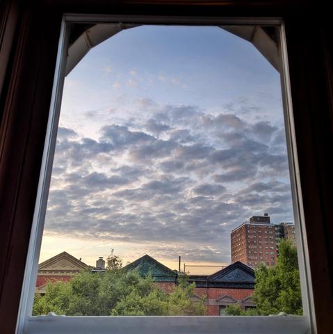 Looking through an arched window fifteen minutes after sunrise the sky fades from palest orange at the horizon to dusty blue up above. A wide band of scattered gray-blue clouds fills the center sky. Pointed roofs of Harlem brownstones with red brickwork are across the street, and a taller apartment building can be seen in the distance.