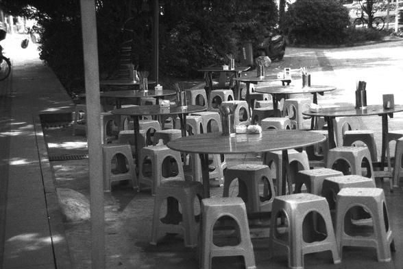 Ilford FP4 Plus 125 (FF)

English alt text: Outdoor dining setup with round tables surrounded by small plastic stools, each table equipped with metal utensil holders, napkin dispensers, and condiments. The area is shaded by trees, with a scooter parked nearby and a quiet sidewalk in the background, suggesting a relaxed street food or casual dining environment.

中文替代文字： 户外餐饮区布置有多张圆桌，周围是小型塑料凳。每张桌上都配有金属餐具筒、纸巾盒和调味品。环境在树荫下显得清凉宁静，旁边有一辆摩托车和人行道，营造出轻松的街头美食氛围。
