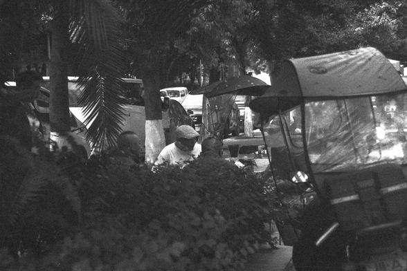 Ilford FP4 Plus 125 (FF)

English alt text: A black-and-white photo of people gathered outdoors around a table, set amidst trees and plants. In the background, vehicles and a rickshaw suggest an urban park setting. The image captures a moment of quiet social interaction, framed by natural and city elements.

中文替代文字： 黑白照片中，人们围坐在户外的一张桌旁，四周有树木和植物。背景中的人力车和车辆暗示这是一个城市公园场景。画面展现了一种城市自然交融中的社交片刻。