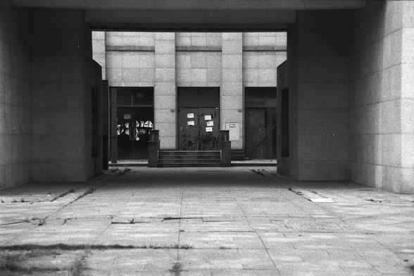Ilford FP4 Plus 125 (FF)

English alt text: A grainy black-and-white photograph of a building entrance composed of large stone blocks. The scene features three doorways, with the center one elevated by several steps and marked by papers or notices. The ground is paved with square stone tiles, some with tufts of grass peeking through, suggesting age and minimal upkeep. The image evokes a quiet, historical atmosphere.

中文替代文字： 一张黑白颗粒感照片，展示了一座由大块石材构成的建筑入口。画面中有三个门廊，中间的门廊有台阶，并贴有纸张或公告。地面由方形石砖铺成，间隙中长有…