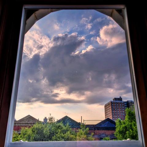 Looking through an arched window two hours after sunrise the blue sky is filled with a large horizontal column of puffy gray and white clouds. Pointed roofs of Harlem brownstones with red brickwork are across the street, and a taller apartment building can be seen in the distance. The green tops of two trees are on the bottom and right.