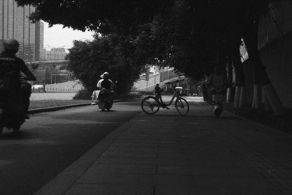 Ilford FP4 Plus 125 (FF)

🚲 English Alt Text

A black and white urban street scene where several people ride bicycles and scooters along a shaded path. A bicycle is parked on the sidewalk, and leafy trees create a canopy overhead. In the background, tall buildings and an overpass suggest a bustling city environment. The image captures a slice of daily life and the active use of personal transport.

🛵 中文替代文字

这是一幅黑白城市街景，多位行人骑着自行车和滑板车在树荫覆盖的小路上行驶。人行道旁停着一辆自行车，郁郁葱葱的树木在头顶形成遮蔽。背景中可见高楼和高架桥，体现了都市生活的繁忙节奏…