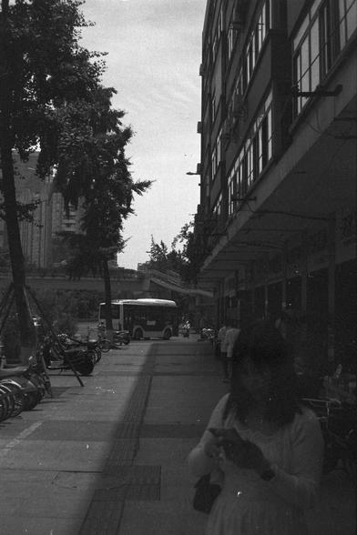 Ilford FP4 Plus 125 (FF)

🚌 English Alt Text

A black and white photo of a city sidewalk with parked bicycles and pedestrians walking past a large building on the right. The building features several windows and mounted air conditioning units. In the background, a bus is partially visible on the street. One person in the foreground looks at their phone while walking. The overall mood feels urban and contemplative.

🚶 中文替代文字

这是一张城市人行道的黑白照片，左侧停放着几辆自行车，行人走过右侧的一栋大楼。建筑外墙有多个窗户和空调机组。背景中隐约可见一辆公交车驶过。前景…