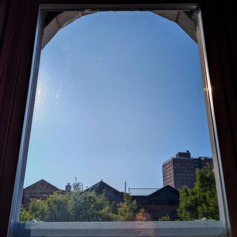 Looking through an arched window one hundred and forty minutes after sunrise the sky is bright blue and cloudkess. Pointed roofs of Harlem brownstones are silhouetted across the street, and a taller apartment building can be seen in the distance. The green tops of two trees are on the bottom and right. 