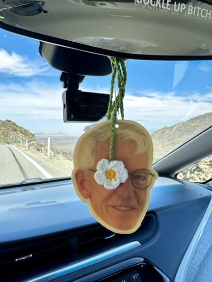 iPhone photo of my air freshener (it’s made from a funny pic of my husband’s face) hanging from the rearview mirror of my car, there’s a white crocheted flower over his right eye, the background features an asphalt mountain road, desert, and open sky with white clouds.