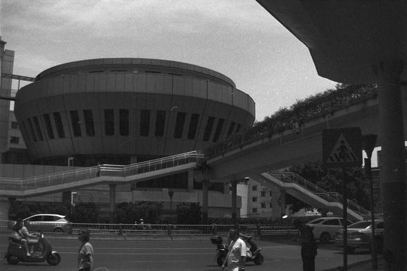 Ilford FP4 Plus 125 (FF)

English alt text: A monochrome photo of a sleek, circular modern building with tall vertical windows. A raised pedestrian walkway curves in front of the structure, featuring railings and a staircase that leads to a busy street below. People walk and ride scooters beneath, near a traffic sign for a pedestrian crossing.

中文替代文字：一张黑白照片展示一座圆形现代建筑，建筑外立面设有高大的竖窗。建筑前是一个弯曲的高架人行道，配有栏杆和通往街道的楼梯。街道上行人和电动车穿梭，还有一个行人过街的交通标志。