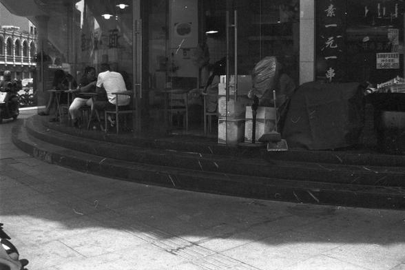 Ilford FP4 Plus 125 (FF)

English alt text: A street-level view of a restaurant with glass windows and doors, showing patrons seated inside. In front of the entrance, steps lead up from the sidewalk, where miscellaneous items such as boxes and cleaning tools are scattered. A motorcycle passes by on the road, while other buildings line the background.

中文替代文字：一张街景照片展示一家小餐厅，玻璃门窗内有顾客正在就坐。餐厅门前的台阶连接人行道，地面上散放着一些杂物，包括纸箱和清洁工具。一辆摩托车从街上驶过，背景中还有其他建筑物。