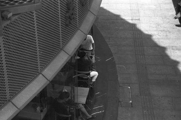 Ilford FP4 Plus 125 (FF)

English alt text: Outdoor seating with three people at separate tables, casually enjoying conversation or activities. The space is set beside a building with a textured metal façade, paved with tiles and illuminated by soft daylight. Shadows stretch across the ground, adding contrast and depth to the urban scene.

中文替代文字：户外座位区，三位顾客分别坐在不同的桌旁，享受交流或各自的活动。场地位于一栋金属镂空外墙的建筑旁，地面铺有瓷砖，阳光柔和洒落，在地面上形成富有层次的阴影，为都市景象增添视觉对比。