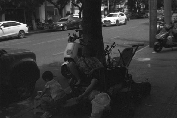 Ilford FP4 Plus 125 (FF)

English alt text: A quiet urban street scene in black and white featuring a person seated on a chair or cart beneath a tree, with a small child nearby. Around them are bags and miscellaneous belongings, including what appears to be a stroller. Scooters and cars are parked along the street in the background, and buildings line both sides, creating a nostalgic and contemplative atmosphere.

中文替代文字：一幅黑白城市街景照片，画面中一位成人坐在树下的椅子或手推车上，旁边有一个小孩。周围散落着一些袋子和随身物品，包括疑似婴儿车。背景中街道两侧停着电动车…
